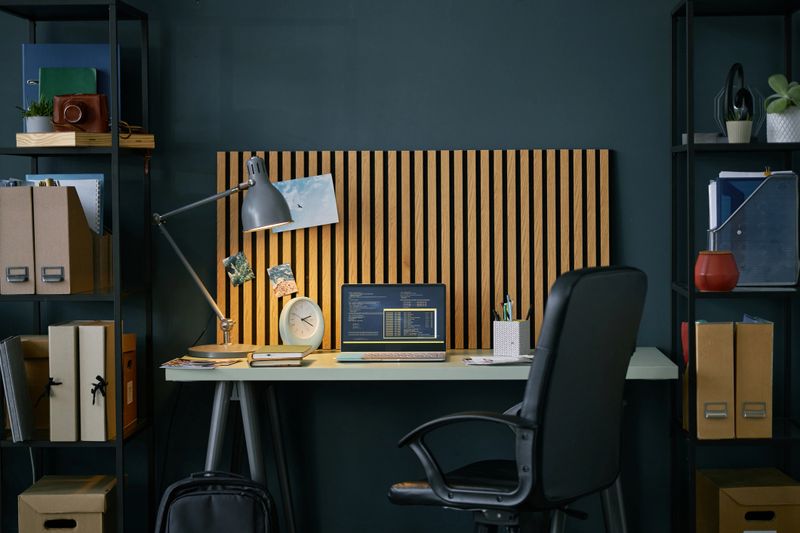 Modern home office setup with a wooden backdrop, a desk lamp, and a laptop on a white table surrounded by black shelves filled with books and storage boxes