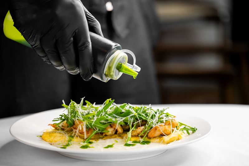 A chef's gloved hand drizzles green sauce over a gourmet dish of grilled chicken, arugula, and grated cheese, highlighting precision, freshness, and fine dining aesthetics.