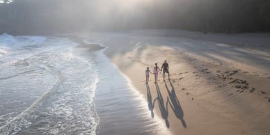 A family of three holding hands and walking on a misty beach at sunrise.