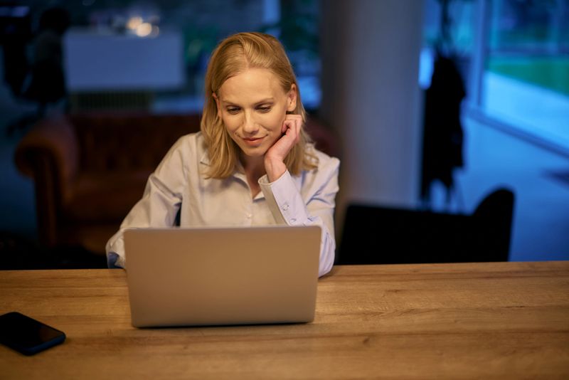 Young businesswoman is sitting at wooden desk in her office at night, working on laptop computer with focused expression, illuminated by warm light