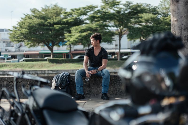 Asian male motorcyclist relaxing after a ride, holding a water bottle and enjoying a quiet moment outdoors. Jacket and gear nearby, parked motorcycle in foreground, modern urban lifestyle.