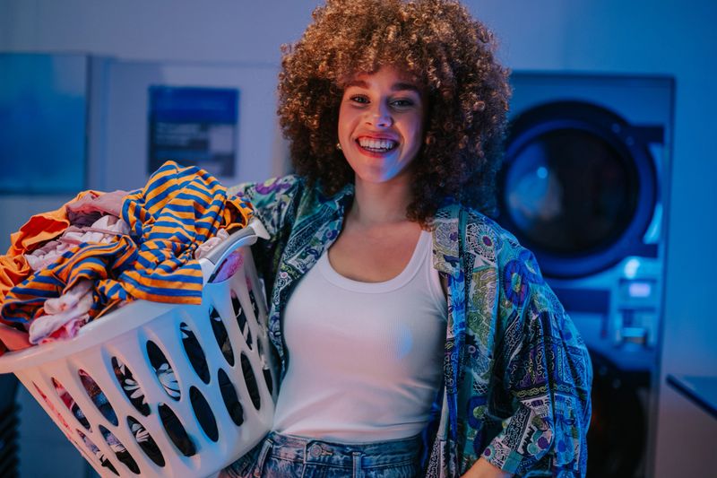 Happy young woman carrying a laundry basket full of clothes in a self-service laundromat, smiling at camera