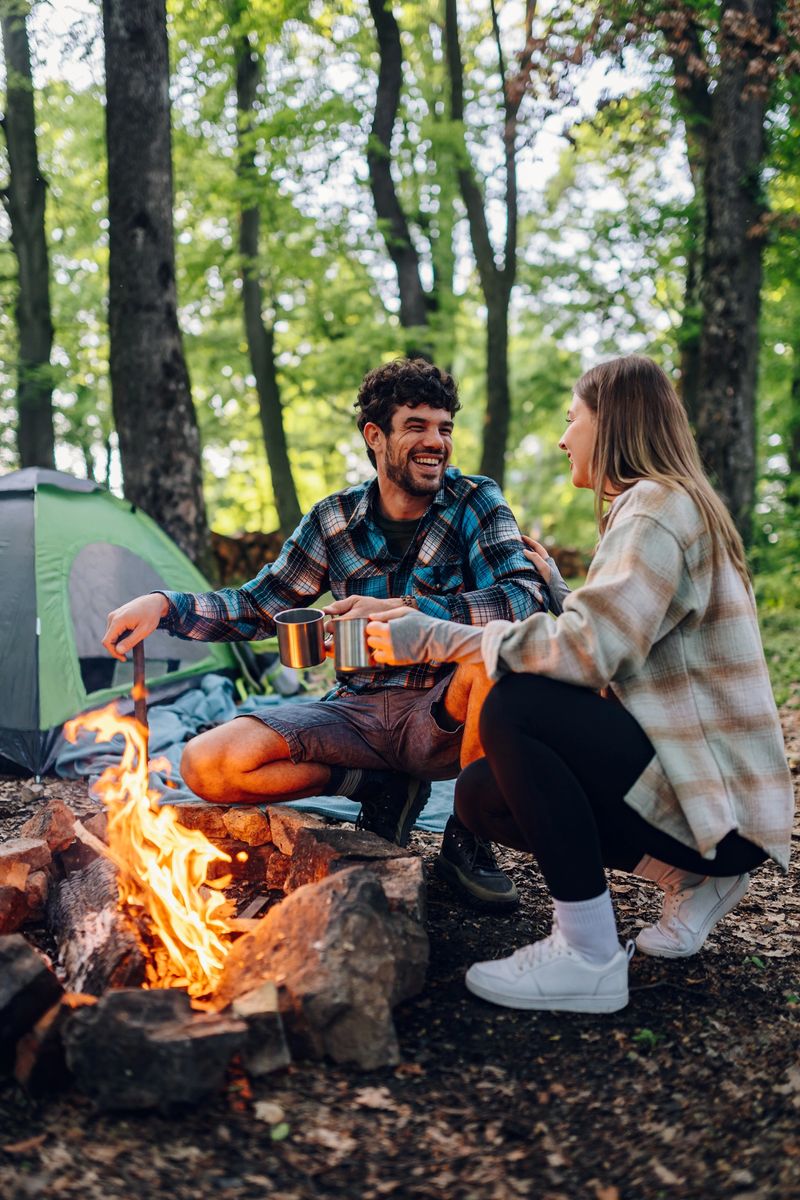 Young couple enjoying a camping trip, sipping coffee by the warm bonfire, sharing stories, and laughing together under the stars