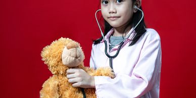 Young girl dressed as a doctor examining a teddy bear with a stethoscope.