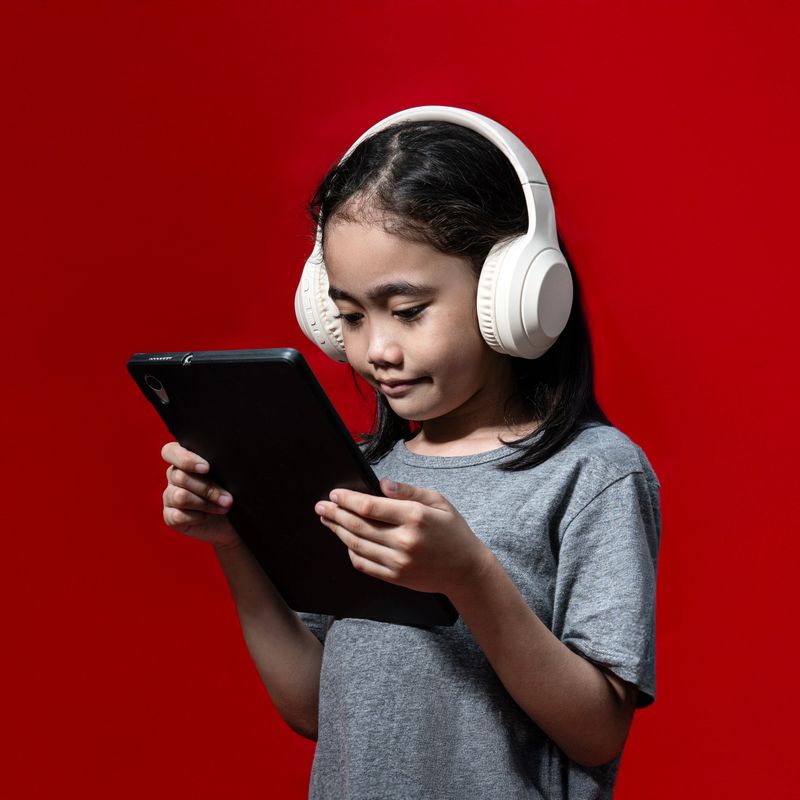 Asian girl wearing large headphones and holding black tablet isolated on red background.