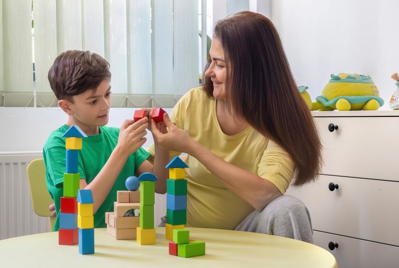 Happy young boy and a woman, possibly his mother or therapist, are playing together with colorful wooden blocks at a table in a bright, cozy home environment. The boy is smiling and engaged, demonstrating focus and curiosity as he explores the tactile shapes and structures. The woman gently supports and encourages him, reinforcing a warm and trusting connection. This scene represents the values of early childhood education, autism therapy, ADHD support, and occupational therapy. It also highlights the importance of sensory learning, emotional bonding, and developmental activities in a nurturing and inclusive setting. Ideal for illustrating topics in child psychology, neurodiversity, home education, and parent-child interaction.