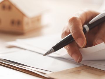 Hand holding a pen writing on paper with a small wooden house model in the background.