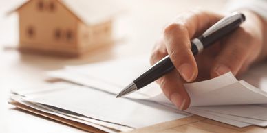 Hand holding a pen over paperwork with a small wooden house model.