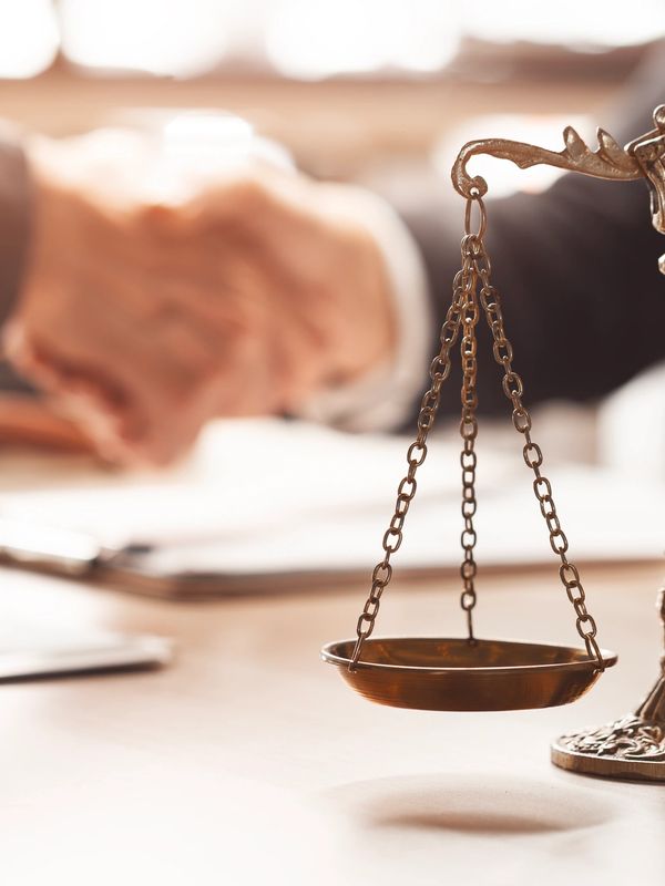 Two people shaking hands behind a justice scale on a desk.