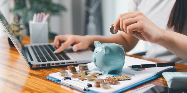 Person saving money by putting coins into a piggy bank while using a laptop.