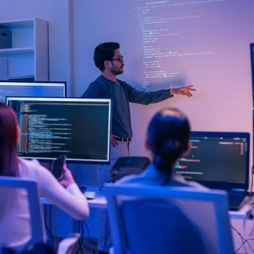 A man presents coding on a projector to a group in a tech setting.