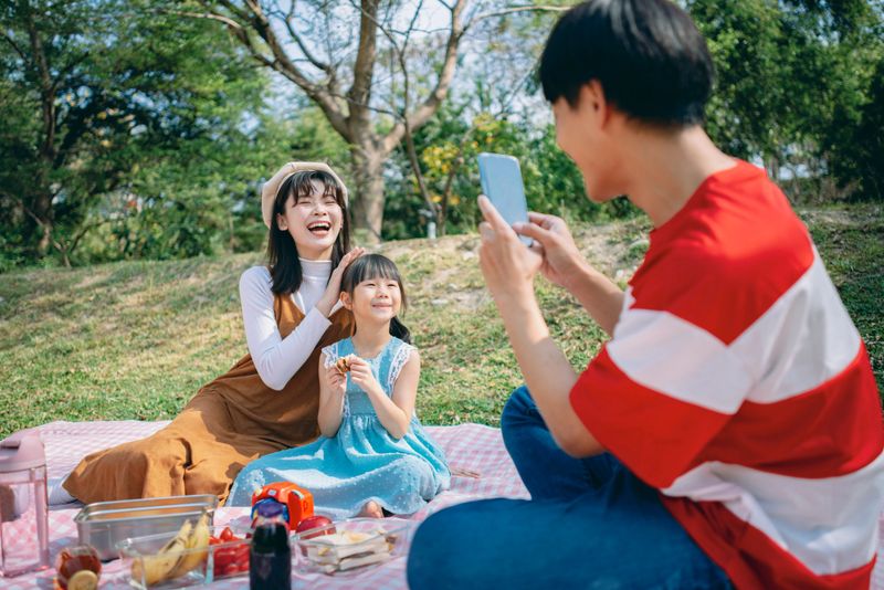 An Asian family enjoys a picnic in the park, creating precious memories together. The father uses a smartphone to record everyday moments and the child’s growth, instantly sharing them on social media with friends and relatives. In the background, rising inflation and financial stress challenge modern parents to strike a balance between work and home. This image captures not only connection and presence, but also how families adapt emotionally and digitally to a changing economic reality.