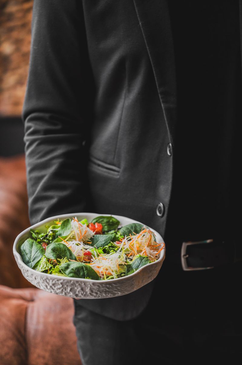 A person in a black suit holds a bowl of fresh salad with spinach, shredded vegetables, and tomatoes. The vibrant colors and textures of the dish contrast with the blurred background