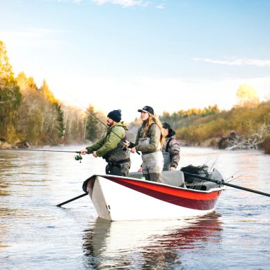 Three people fishing from a boat on a calm river surrounded by autumn trees.