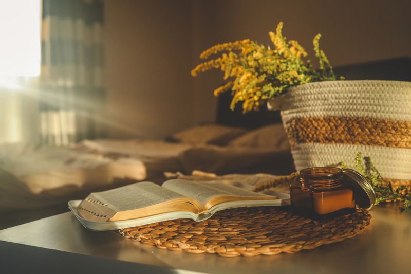 Cozy home scene with a burning candle in an amber glass jar, open book, and yellow wildflowers on a woven placemat in warm natural sunlight.