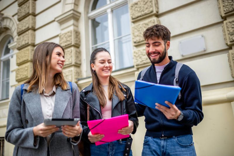 Three students stand together outdoors, holding books and literature, engaging in a lively conversation filled with laughter. Dressed casually and carrying backpacks.