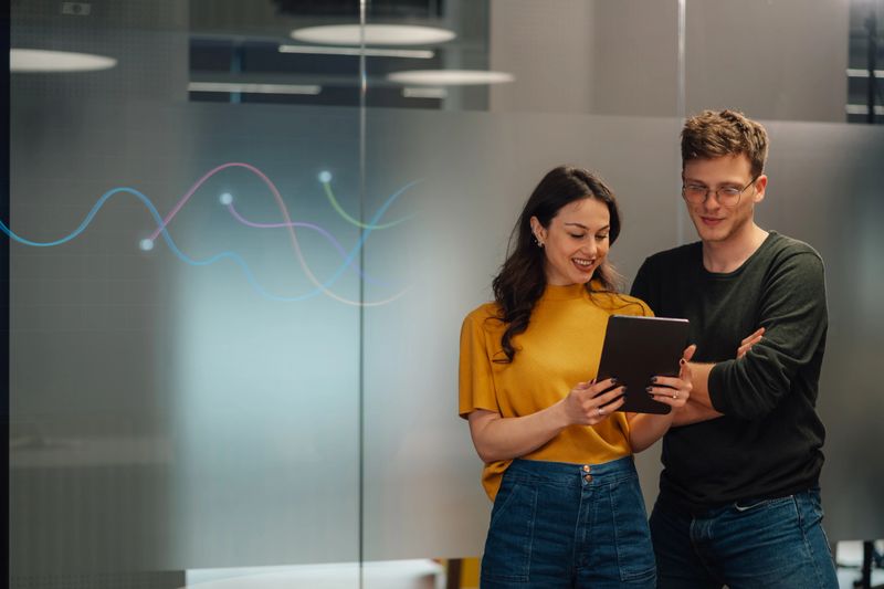 Two young professionals are smiling while reviewing data on a tablet in a modern office, suggesting a collaborative and positive work environment