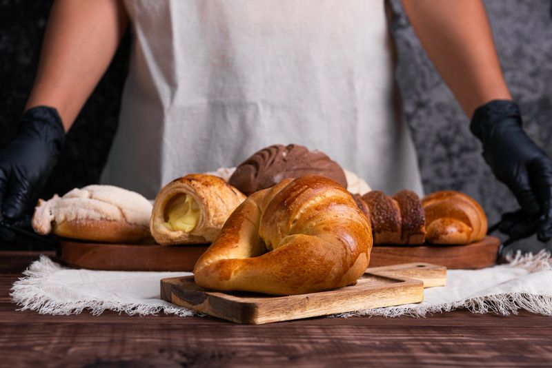 A baker displays an assortment of freshly baked goods, including a golden croissant, concha bread, and cream-filled pastries.