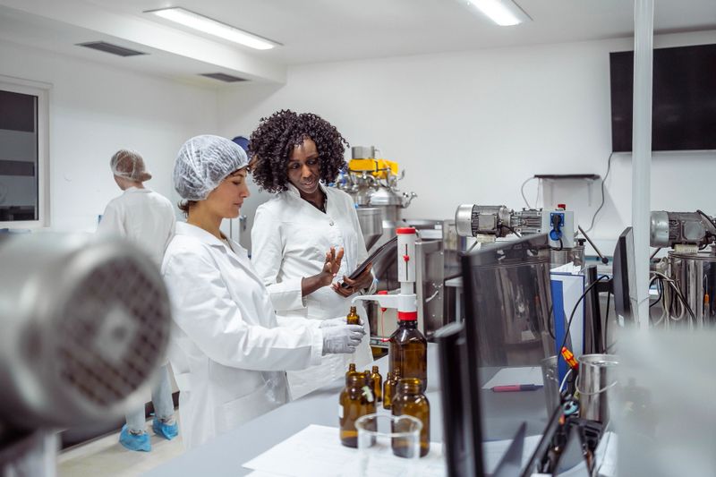 Close-up view of mid-aged diverse female scientists in protective gear examining pharmaceutical samples while performing quality control procedures in sterile laboratory environment.