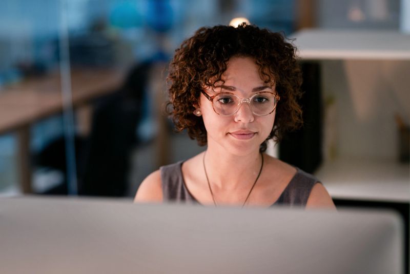 A young woman with curly hair and glasses concentrates intently while using a computer in a well-lit modern office environment. The atmosphere conveys a professional and productive workspace.