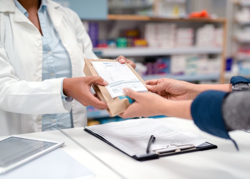 A pharmacist hands a package of medication to a customer across the counter, symbolizing trust and care. The scene portrays professionalism and customer interaction in a healthcare setting. Shelves of medicines in the background and visible documents on the counter emphasize the clinical environment, highlighting the importance of accuracy and guidance in pharmacy services.