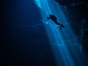 Scuba diver illuminated by shafts of light underwater in a cave.