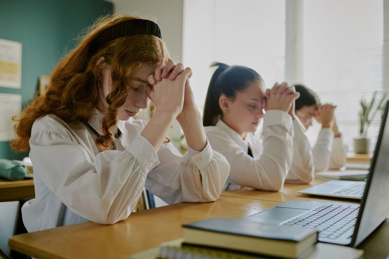 Group of students in school uniforms praying with closed eyes and folded hands while seated at desk in classroom environment