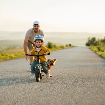 Child learning to ride a bike with father and dog nearby.