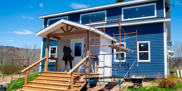 Worker constructing a porch on a blue house under a clear sky.