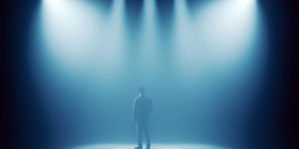A lone figure stands illuminated by multiple spotlights in a dark, foggy space.