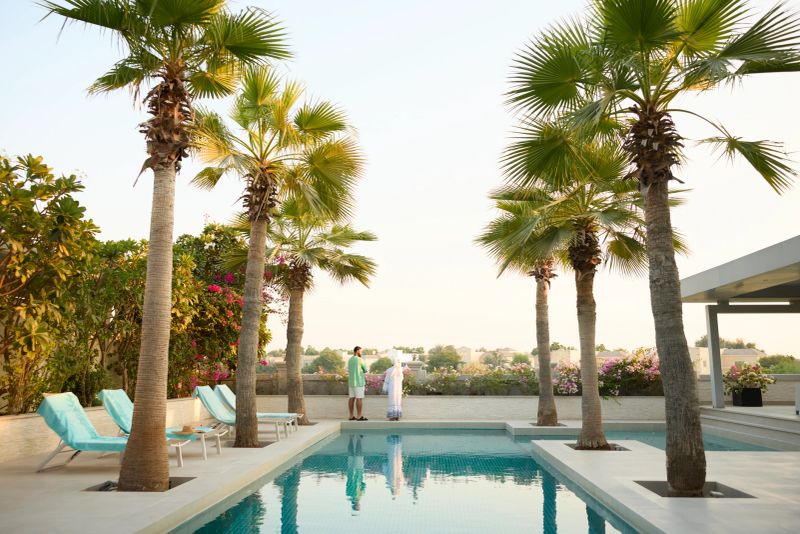 Middle eastern couple spending leisure time talking with each other. Modern swimming pool in back area of villa against clear sky. Palm trees can be seen on poolside.