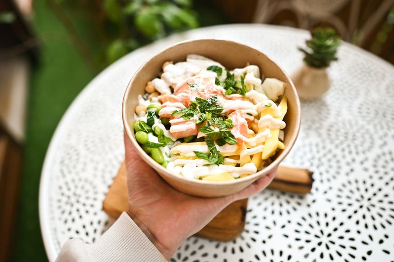 Hand holding a colorful and healthy poke bowl with salmon, mango, edamame, and a creamy sauce
