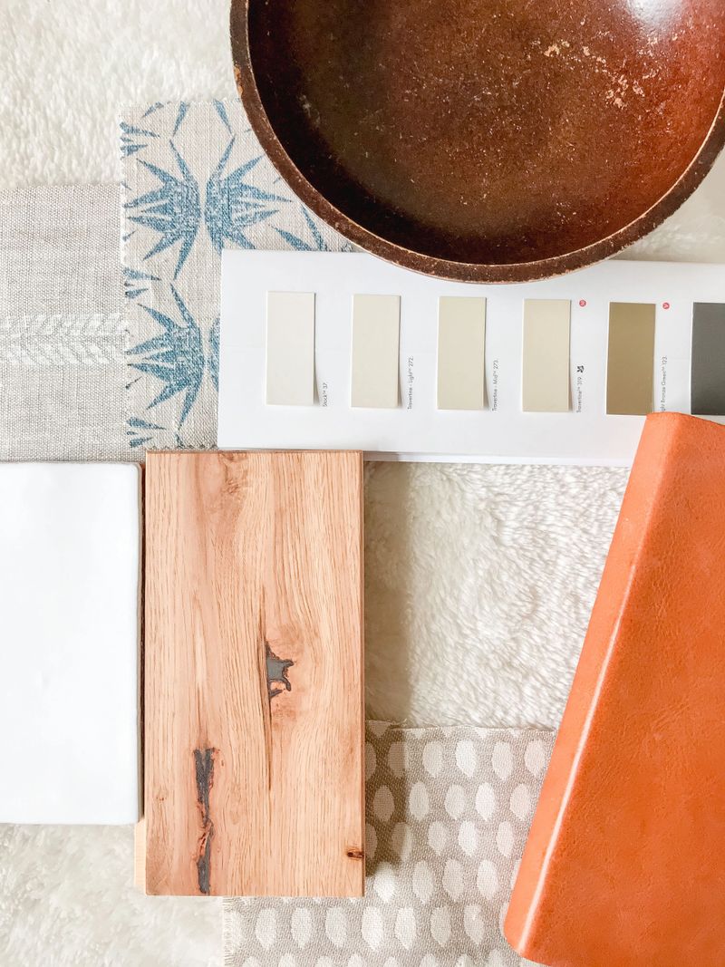 An interior design materials including fabric swatches, a wooden plank, a color palette, and a ceramic bowl arranged on a textured surface.