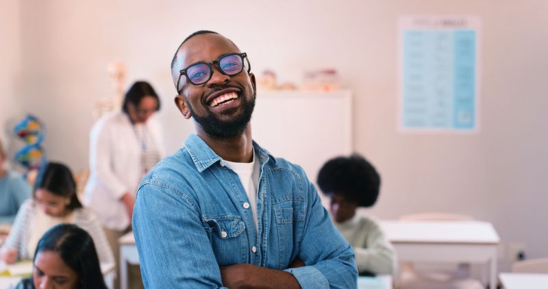 A cheerful, confident instructor stands at the forefront of a bright and lively classroom. The environment reflects engagement and positive learning, with students immersed in teamwork and discussions. Representation of learning and mentoring.
