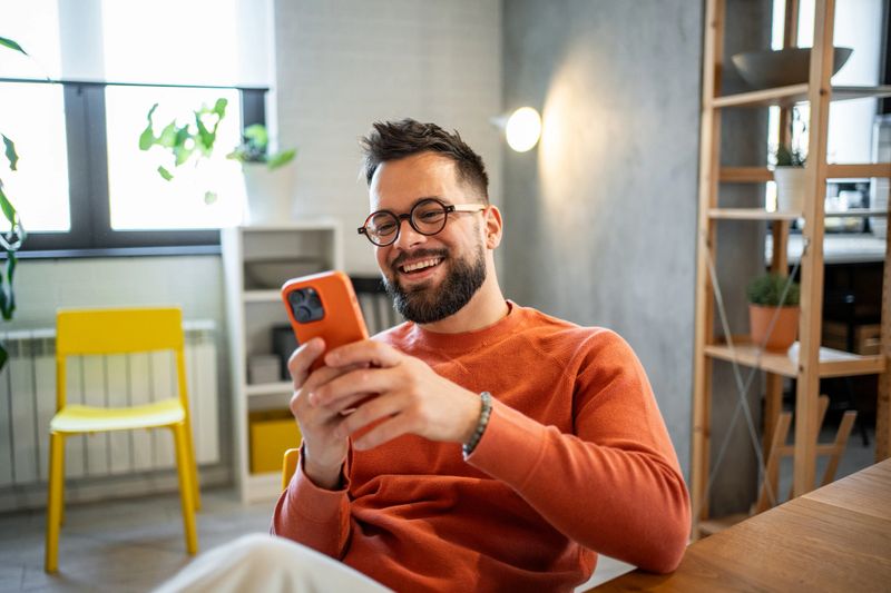 Young adult man with eyeglasses and a beard laughing while using his mobile phone, sitting comfortably in his modern apartment, enjoying a moment of joy and connection