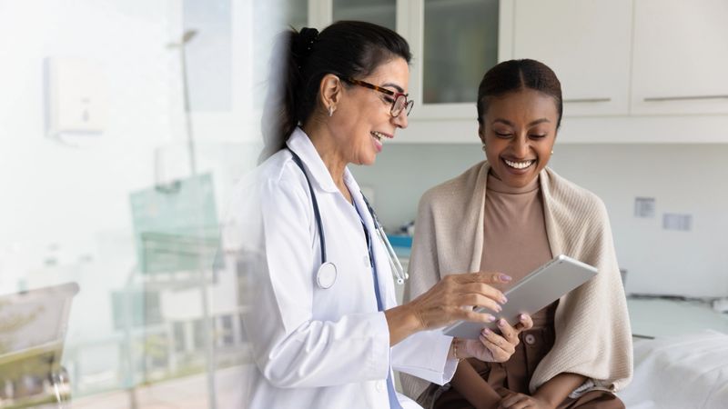 Happy senior practitioner woman and cheerful young African patient woman watching examination medical report on tablet computer, using digital technology, laughing