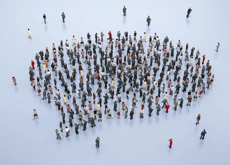 A large group of people dressed in formal and casual attire stand together, forming a unique shape on a smooth floor in a city setting, engaging in discussion or networking.