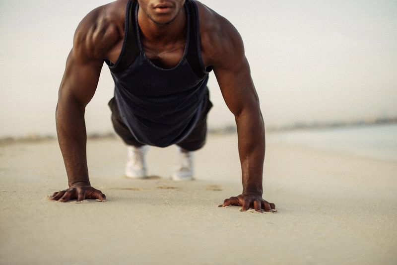 A close-up of a man's torso and arms as he performs push-ups on a beach. The exercise scene captures strength, determination, and fitness against a sandy, outdoors landscape, creating a dynamic and inspiring atmosphere for a healthy lifestyle.