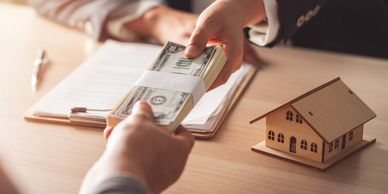 Hands exchanging a stack of cash beside a small rental house model on a desk.