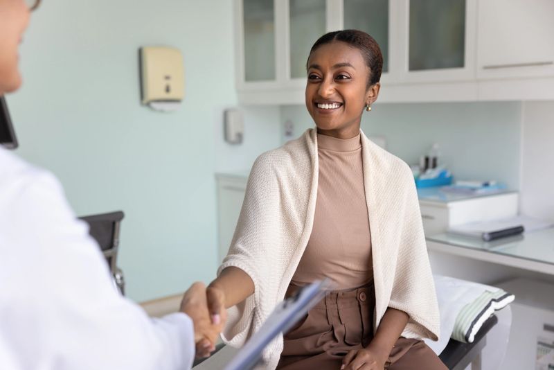 Positive satisfied healthy young African patient giving handshake to female doctor with gratitude, happy smile, sitting on clinic examination couch, thanking medical professional, shaking hand