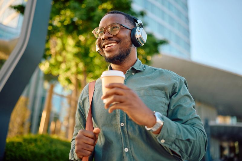 Happy african american businessman is enjoying a coffee break while listening to music with his headphones, walking outside modern office buildings