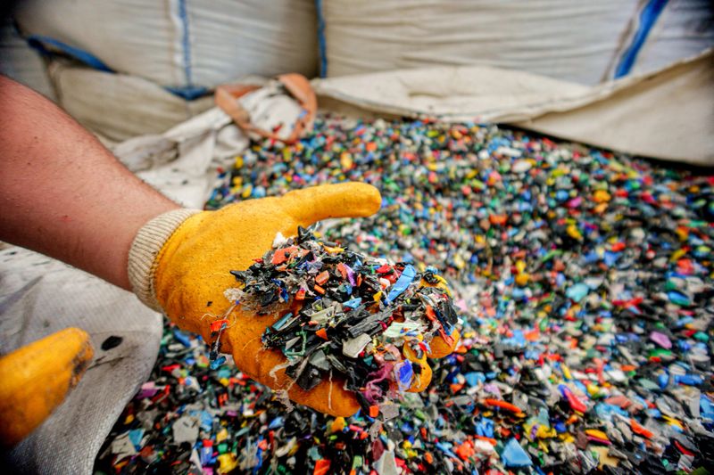 Close-up image of a worker wearing yellow protective gloves holding a handful of multi color recycled plastic pellets. The granules are used as raw materials in plastic manufacturing and injection molding processes. The image represents sustainability, recycling, and industrial production.
