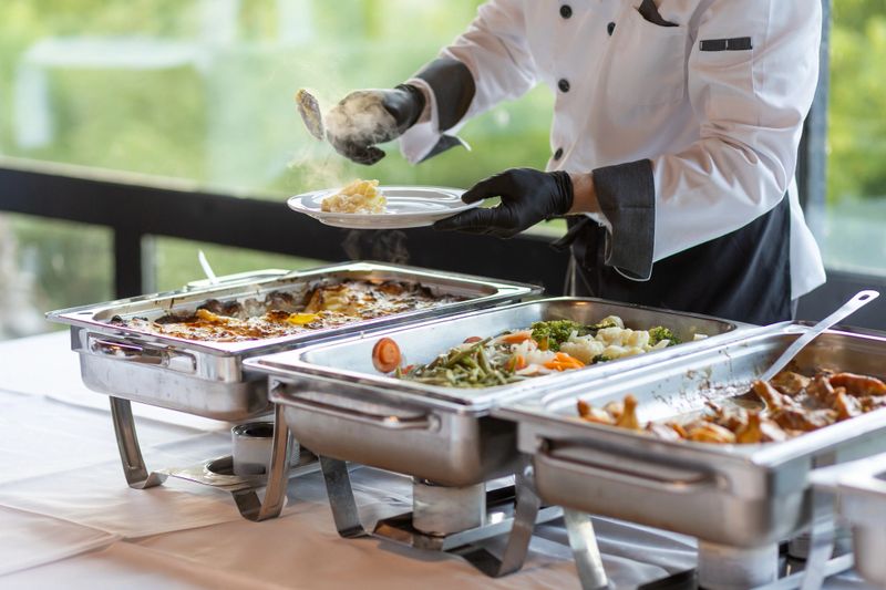 A chef in a black and white uniform expertly plates portions of food from elegant buffet trays. The setting features a bright, contemporary atmosphere with greenery in view.