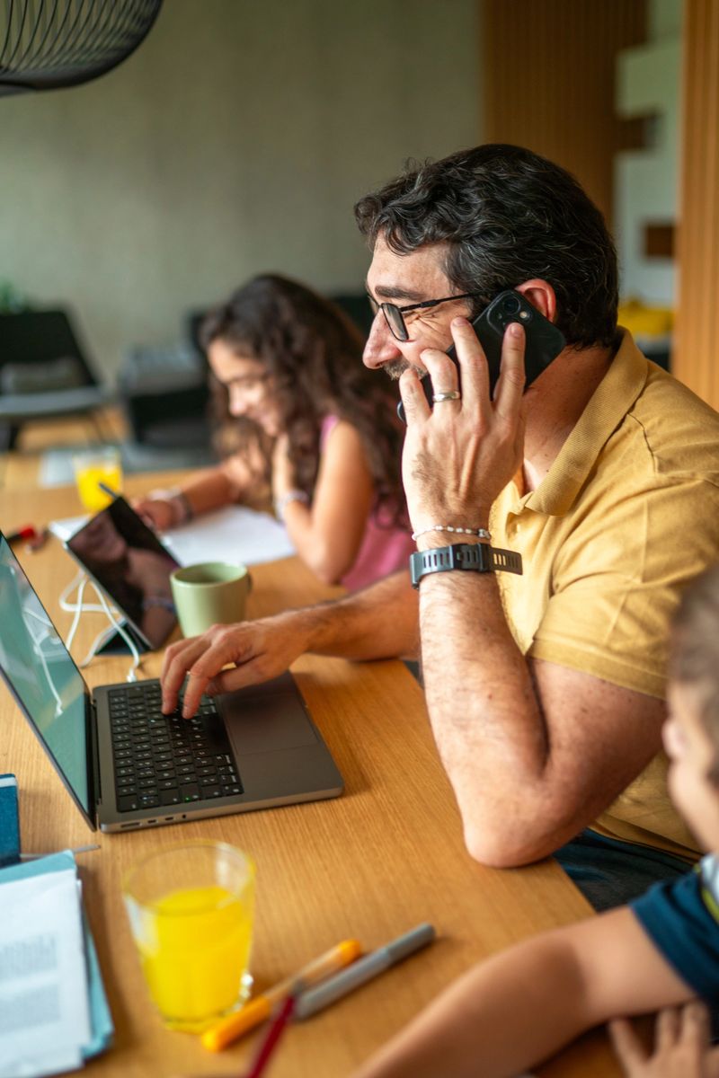 Mid adult Hispanic father multitasks as he works on a laptop while assisting his son and daughter with homework. Seated in a casual domestic dining room, he wears a casual shirt and uses a smartphone, creating a supportive family space.
