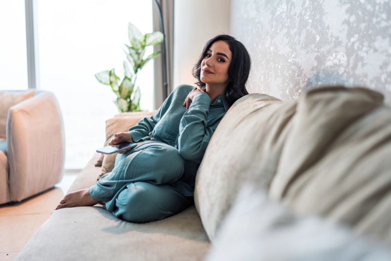 Young Arabic woman in comfortable lounge wear enjoys a peaceful moment on a plush sofa in a stylish, modern home. Natural light and greenery enhance the serene atmosphere.