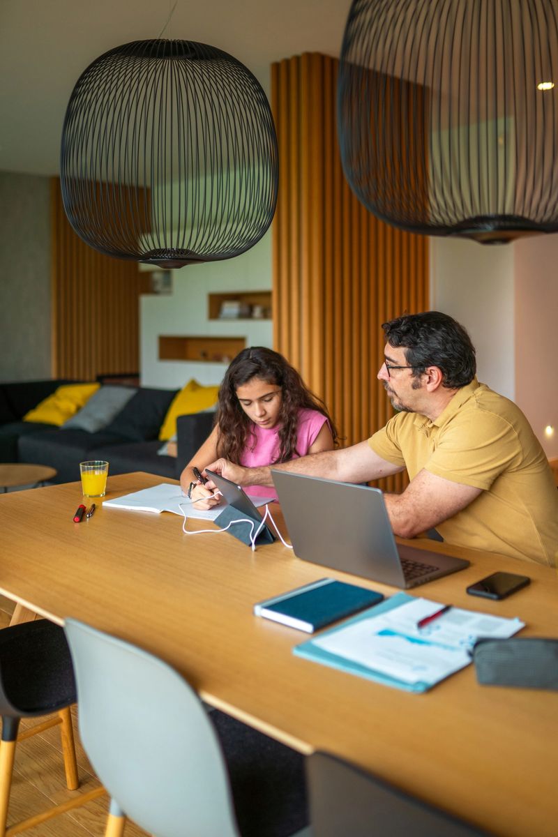Mid adult Hispanic father balances remote work while helping his daughter with homework in a domestic dining room. Both are casually dressed; he uses a laptop while she writes in a notebook. Bright, inviting space with modern lighting and comfortable seating.