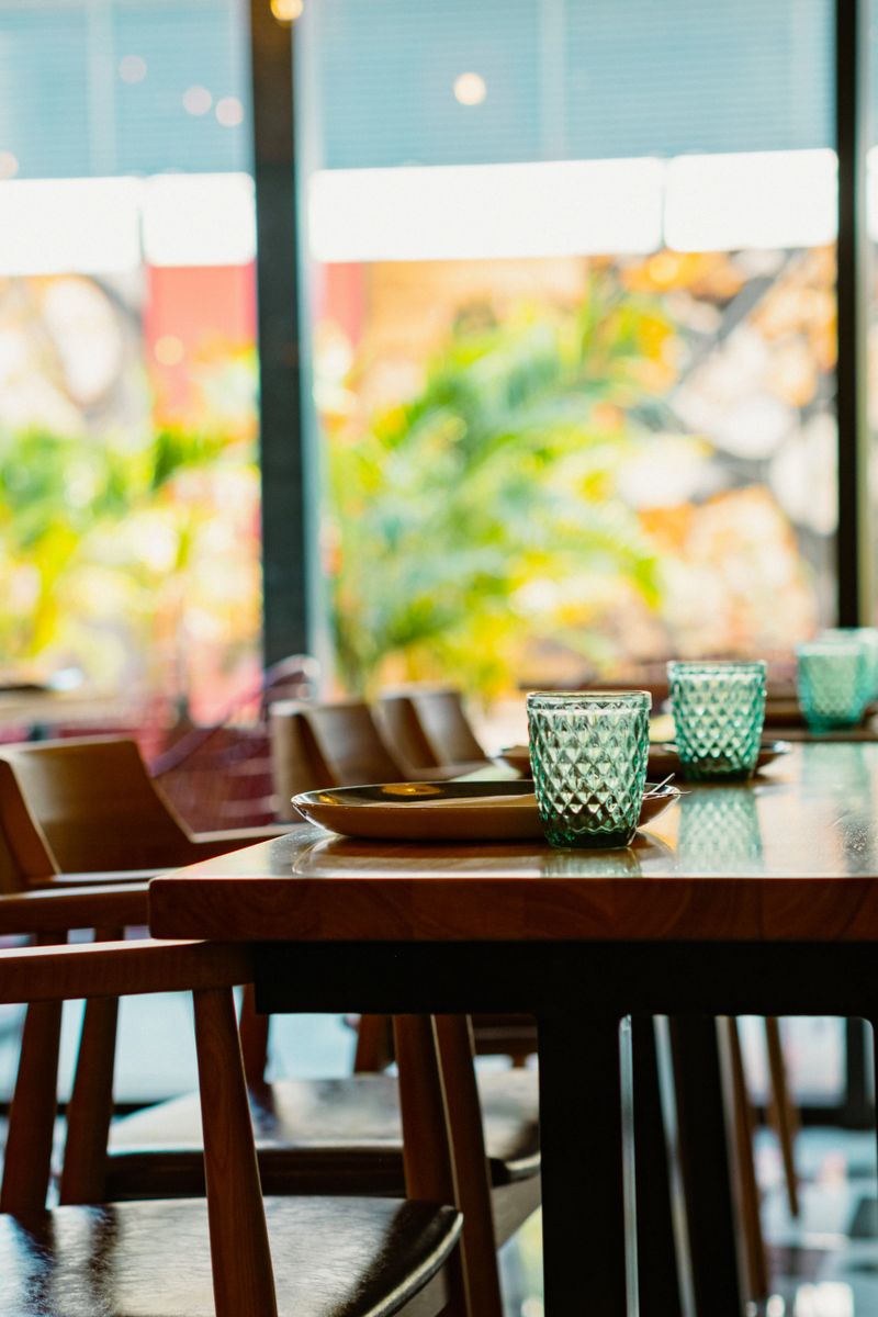 Ready for guests. A long wooden communal table, set with stylish plates and turquoise glasses, basks in bright sunlight, creating an inviting atmosphere for shared meals.
