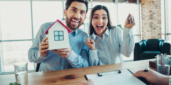 Excited couple holding a house model and keys in a bright office.