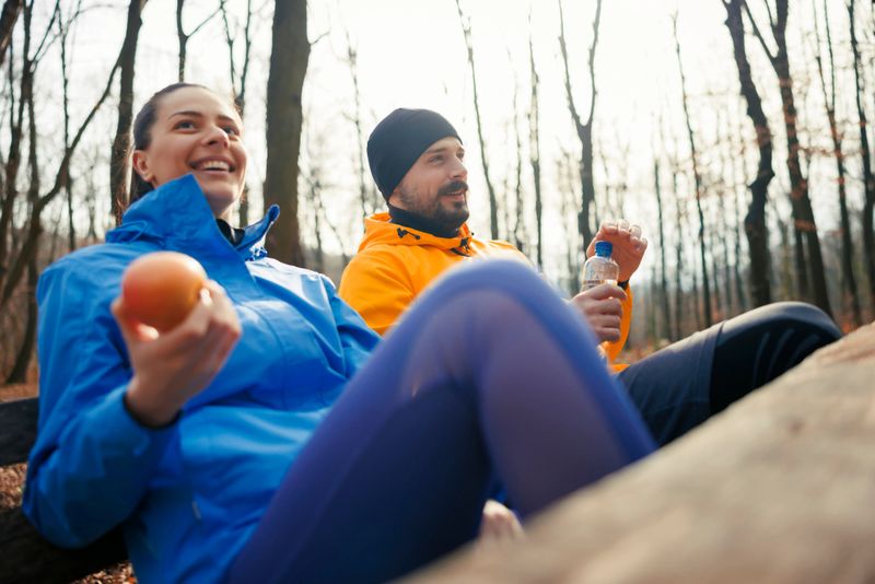Active fit young couple in sportswear taking a break while jogging in nature, sitting on a bench, eating fruits and drinking water