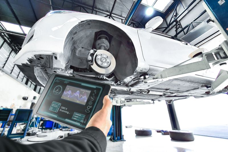 Diagnostic automotive engine system problems, An automotive engineer uses a OBD diagnostic tablet to scan a white car elevated on a lift in a bright service garage.